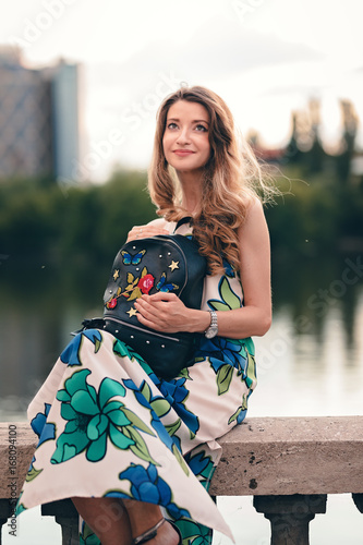 Young blonde girl sitting on a concrete handrail. Looking at the sky and enjoying the pleasant atmosphere. Holding  a stylish designer bag on hand. Long curled hair flowing with wind. photo