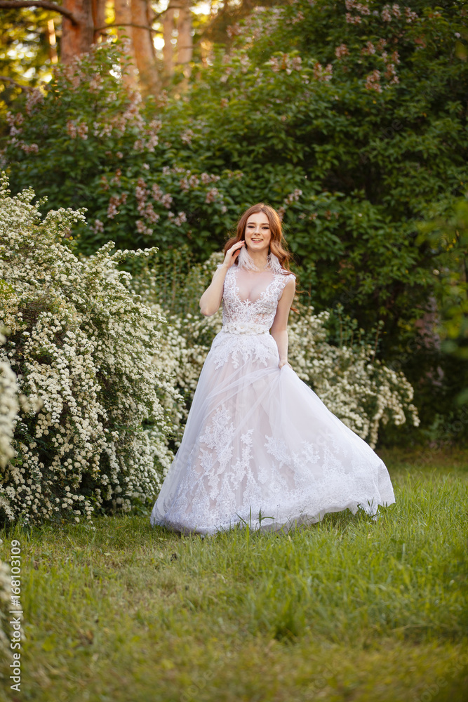 Beautiful redhead Bride in blooming garden playing with her dress. Portrait outdoor in sunset light. Pretty young caucasian redhead girl