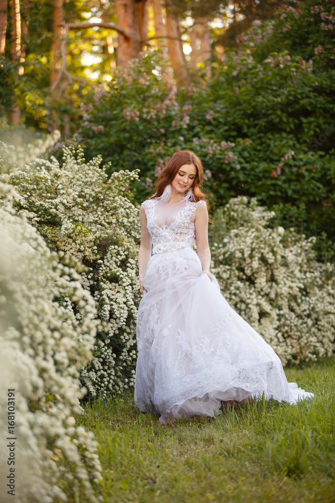 Beautiful redhead Bride in blooming garden playing with her dress. Portrait outdoor in sunset light. Pretty young caucasian redhead girl