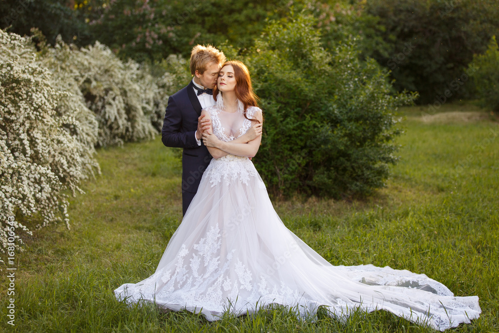 Young Bride and Groom couple in a blooming garden. Tender holding each other. Spring wedding. Redhead girl with long hairs. Young family outdoor image near blooming bush of spirea. Love and tenderness