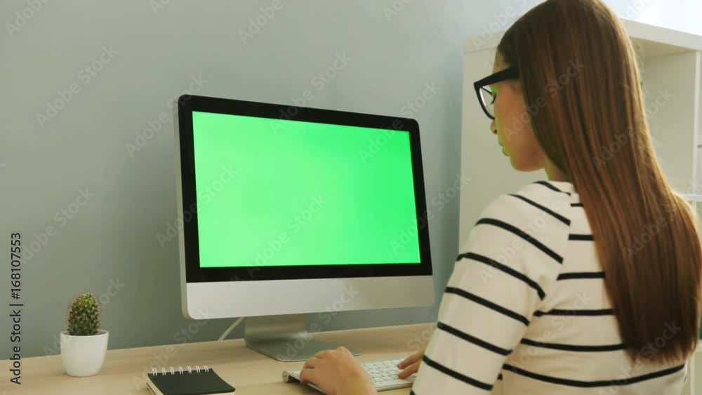 Portrait of young woman in the glasses and casual shirt working on the computer with green screen in the office. Chroma key. View from the back. Portrait of young woman in the glasses and casual shirt working on the computer with green screen in the office. Chroma key. View from the back.