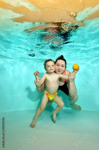 Little boy in diaper with my mother standing underwater at the bottom of the pool, posing and looking at me. Portrait. Shooting under the water surface. Vertical orientation