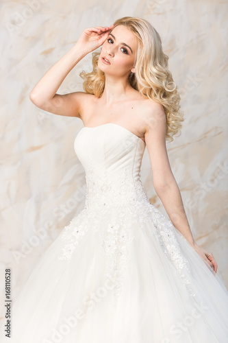fashionable gown, beautiful blonde model, bride hairstyle and makeup concept - young sensual lady in white wedding festive dress, standing indoors on light background, romantic slender woman posing