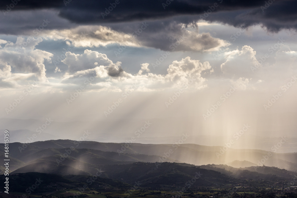 Spectacular view of sun rays and rain over some mountains and hills, benath a moody and cloudy sky