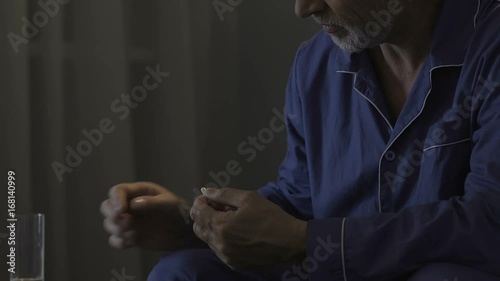 Aged man sitting on edge of bed in darkness, taking medicine, sleeping pill photo