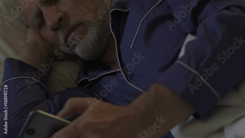 Aged man lying in bed awake at night scrolling mobile phone, problems with sleep photo