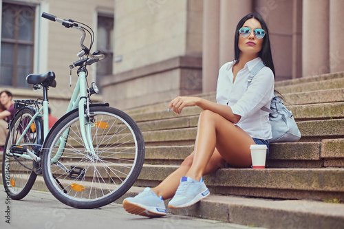 Female sits on a step with city bicycle on background.