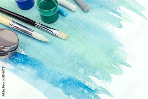 various paintbrushes, paints and pastel chalks on green blue watercolor background