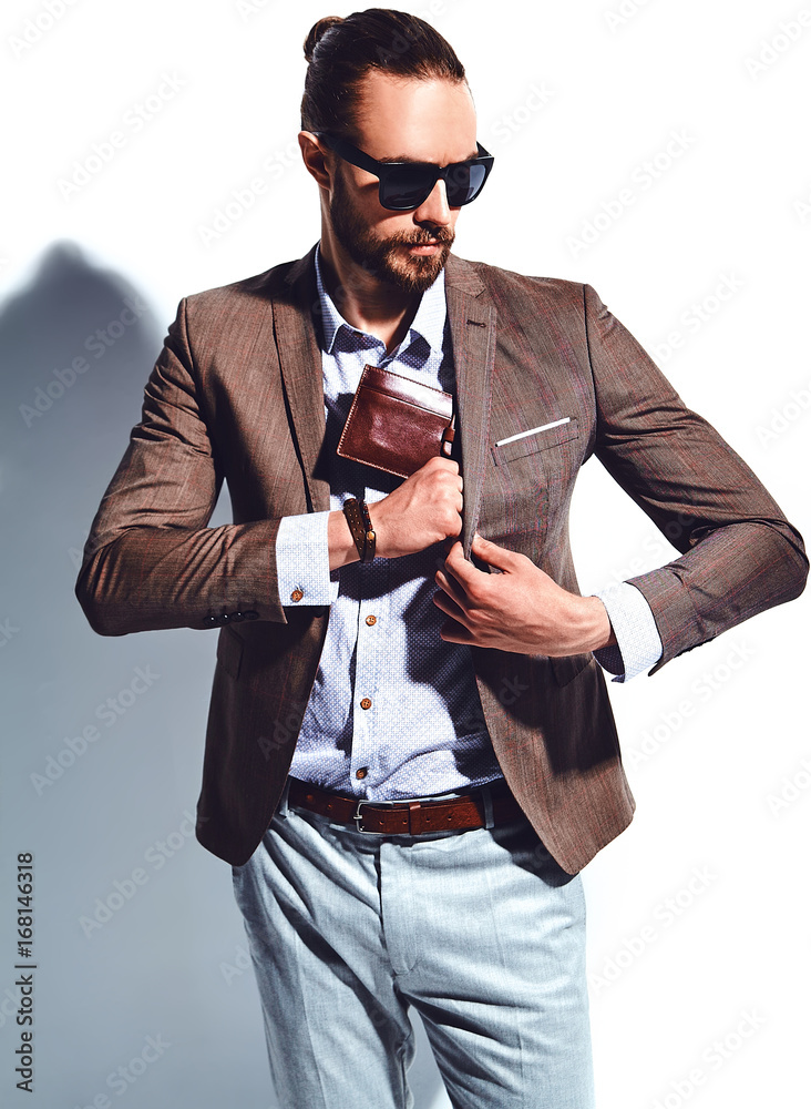 portrait of handsome fashion stylish hipster businessman model dressed in  elegant brown suit in sunglasses posing near white wall in studio. Pulls  out or put his leather wallet into jacket pocket Stock
