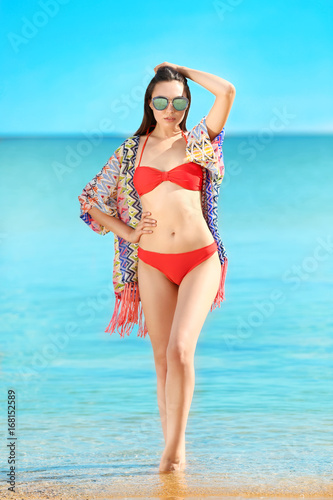 Beautiful young woman in bikini near sea on summer day