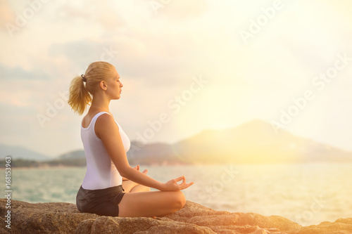 Young woman is practicing yoga on a stone at tropical sea sunrise background.
