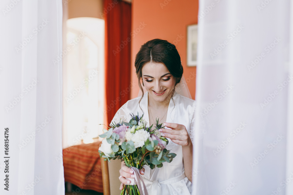 Bride brunette in veil and white satin dressing gown sits by the window Bride brunette in veil and white satin dressing gown sits by the window