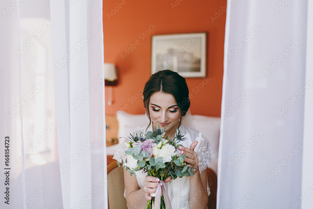 Bride brunette in veil and white satin dressing gown sits by the window Bride brunette in veil and white satin dressing gown sits by the window