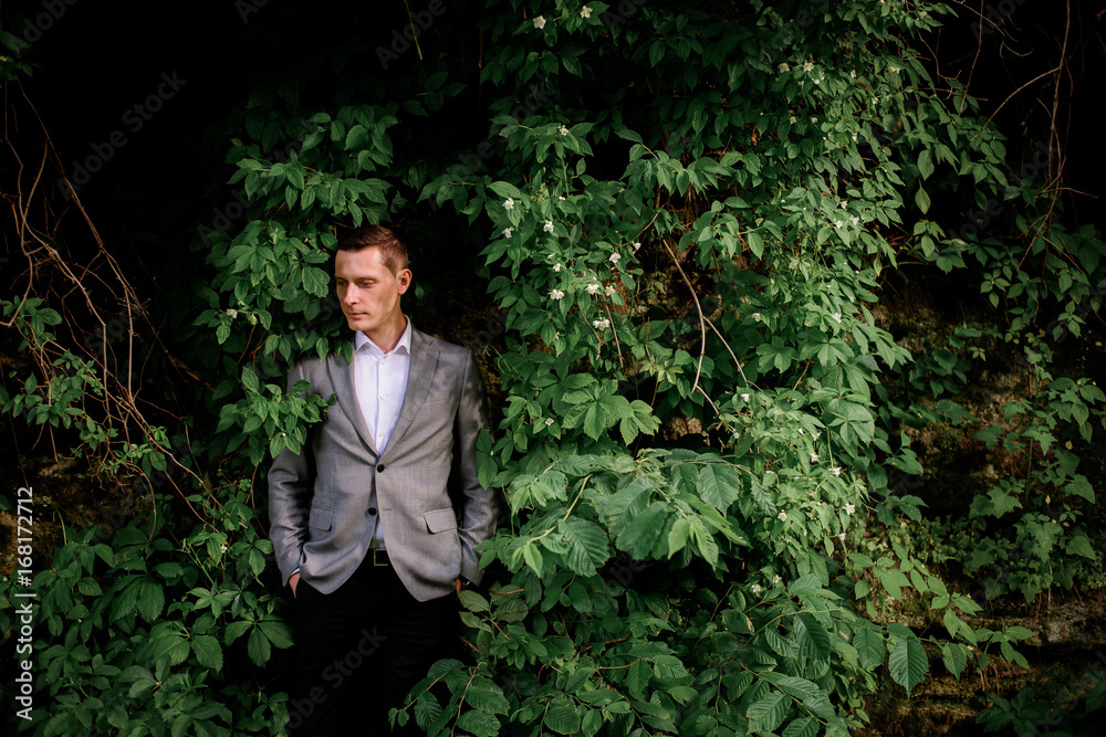 Thoughtful groom stands before a wall with greenery Thoughtful groom stands before a wall with greenery