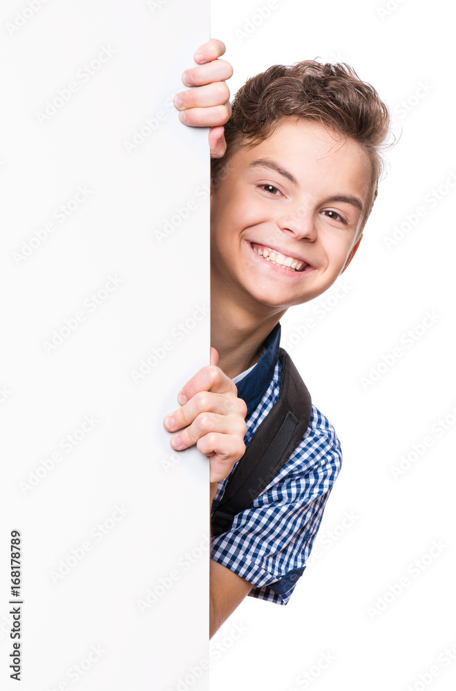 Portrait of handsome teen boy posing behind white panel isolated on white background. Teenager child holding empty billboard. Young student with school bag peeping behind blank board. Portrait of handsome teen boy posing behind white panel isolated on white background. Teenager child holding empty billboard. Young student with school bag peeping behind blank board.