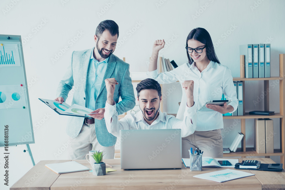 Yes! We did it! Success and team work concept. Team of three business partners with raised up hands with fists in light modern work station, celebrating the growth of their company Yes! We did it! Success and team work concept. Team of three business partners with raised up hands with fists in light modern work station, celebrating the growth of their company