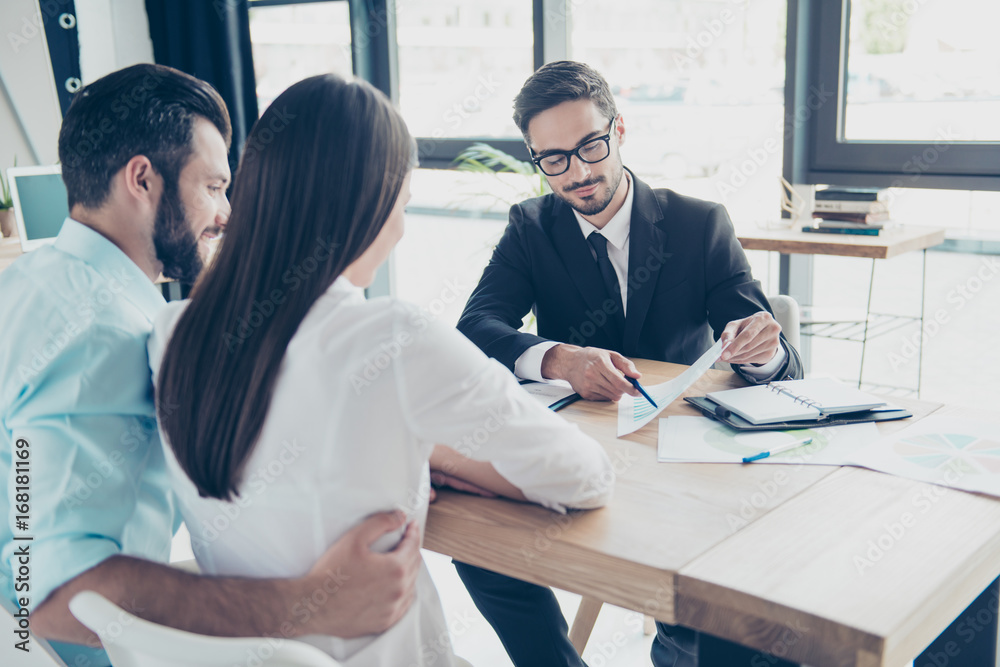 Rear shot of young family, making a deal with lawyer about their business. Men is showing the persentage of a credit of bank, all are well dressed, focused, smiling, sitting in modern office Rear shot of young family, making a deal with lawyer about their business. Men is showing the persentage of a credit of bank, all are well dressed, focused, smiling, sitting in modern office