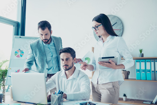 Two colleagues are standing next to the new one, sitting, looking at the laptop screen. Help, unity, tteam work concept, all in formal wear, in modern nice office