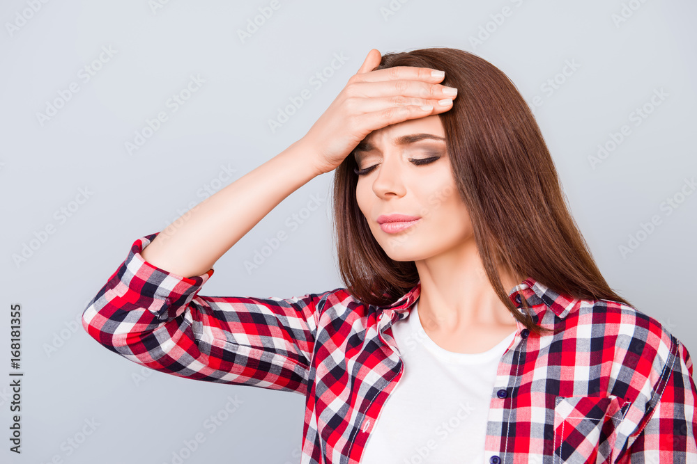 Tired sick pretty brown haired young lady with sad grimace. She is holding the forehead, wearing the checkered shirt, standing on pure background