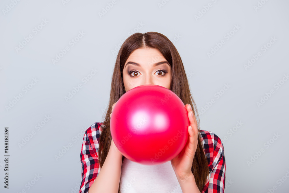 Young amazed brown-haired girl is blowing red balloon for birthday party and looking at the camera on a grey background Young amazed brown-haired girl is blowing red balloon for birthday party and looking at the camera on a grey background