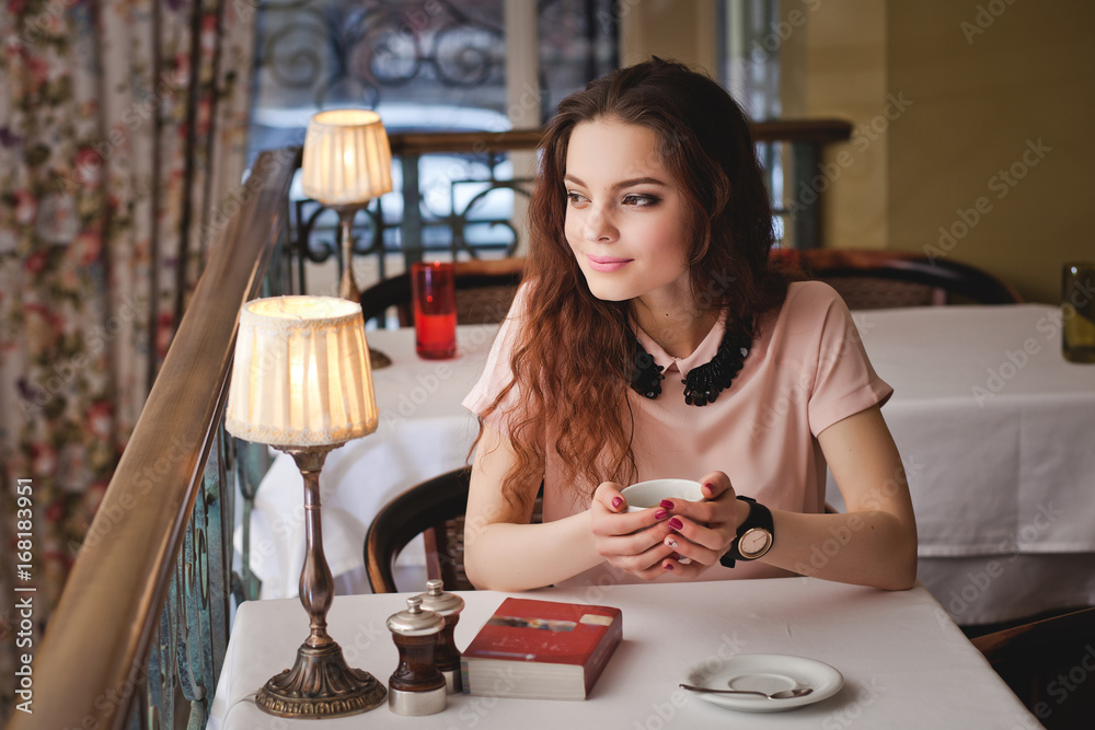 Beautiful young girl sitting alone in a cafe, drinks coffe and has a book