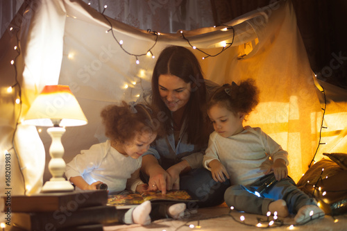 Reading and family games in children's tent. Mother and two twins daughters with books and flashlight before going to bed.