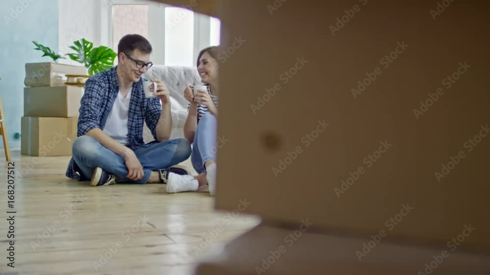 PAN with slow motion of happy young man in glasses and his beautiful girlfriend sitting on wooden floor on new apartment and chatting while drinking coffee PAN with slow motion of happy young man in glasses and his beautiful girlfriend sitting on wooden floor on new apartment and chatting while drinking coffee