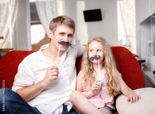 Joyful father and daughter with artificial mustache while sitting togheter on red chair at home.