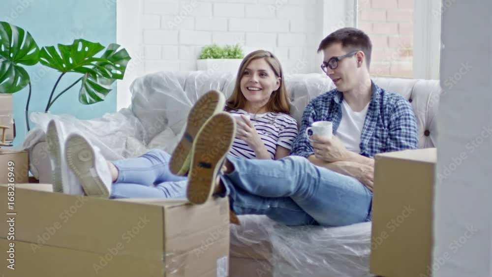 Slow motion of young happy couple sitting on sofa with their legs on cardboard box and chatting while drinking coffee Slow motion of young happy couple sitting on sofa with their legs on cardboard box and chatting while drinking coffee
