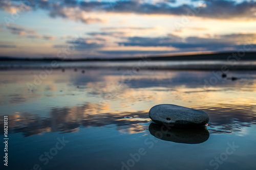 Solitary pebble on wet sand reflecting sunset sky colours.