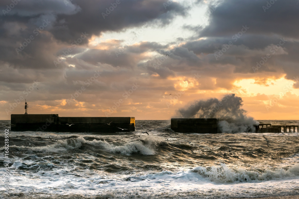 Big wave crashes over Hastings' breakwater under winter stormy sky at sunset