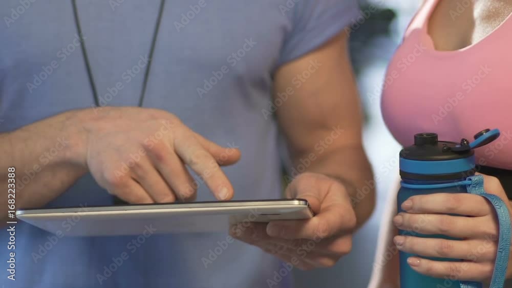 Gym trainer showing schedule on tablet to girl with sports water bottle in hands Gym trainer showing schedule on tablet to girl with sports water bottle in hands
