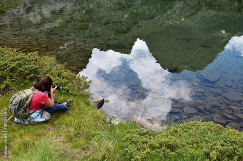 Fototapeta Naklejka Na Ścianę i Meble -  Mature woman takes pictures of a mountain lake with her digital camera, mountain peak reflection seen in the limpid water