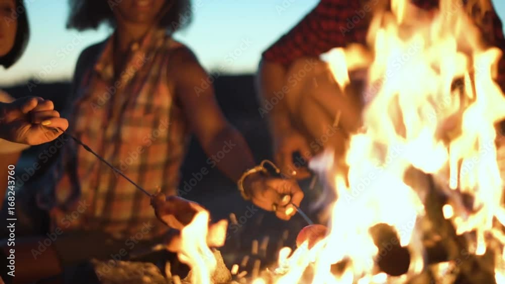 Crop view of people sitting around campfire and grilling sausages on sticks in twilight.