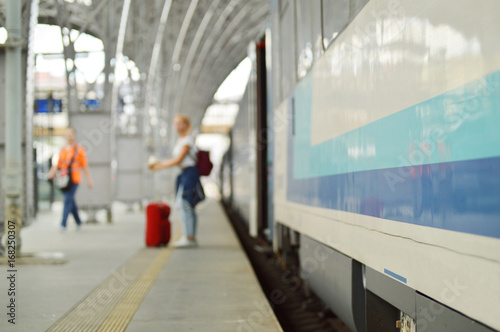 blue and grey train with metal construction, platform and person with suitcase in the background, partly blurred