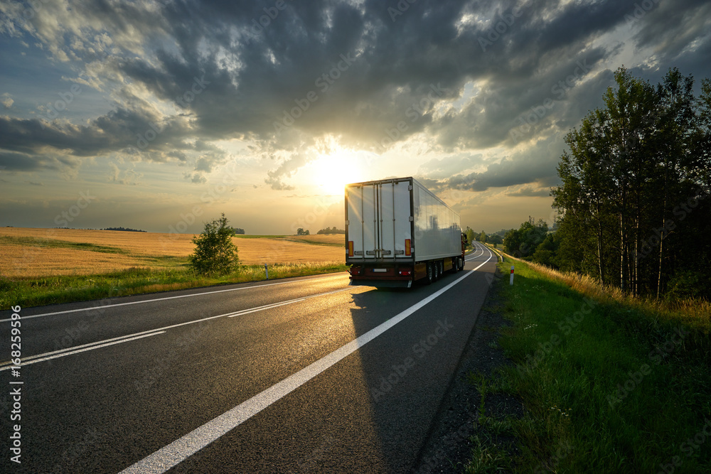 Truck driving on the asphalt road along a field with golden grain in rural landscape at sunset with dark clouds