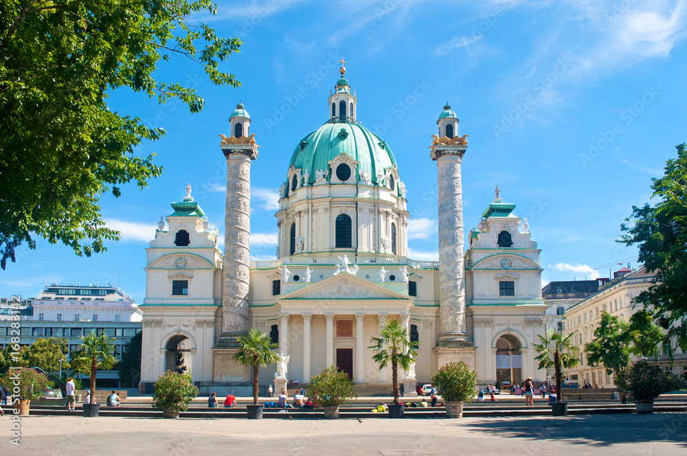 Facade of Karlskirche (St. Charles's Church) against blue sky and several white clouds on a hot summer day. Several pots with palm trees and bushes standing on the city square. Vienna, Austria
