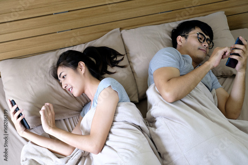 Young happy couple in bed busy with mobile phones separately, A man plays on a smartphone for business and his wife is chatting with her friend on social media i, relax time ,soft focus background