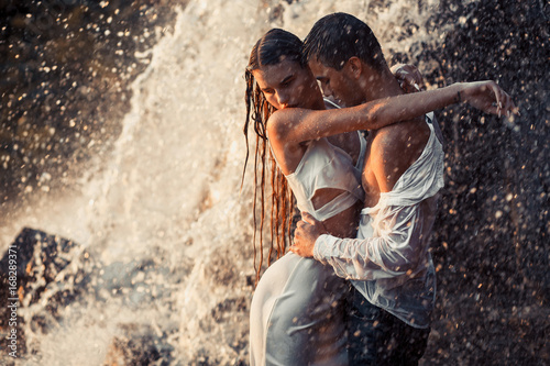 Young enamored couple hugs under spray of waterfall.