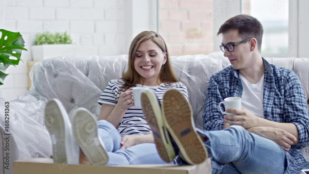 Slow motion of young woman and man sitting on sofa covered in plastic wrap, chatting and drinking coffee in new flat Slow motion of young woman and man sitting on sofa covered in plastic wrap, chatting and drinking coffee in new flat
