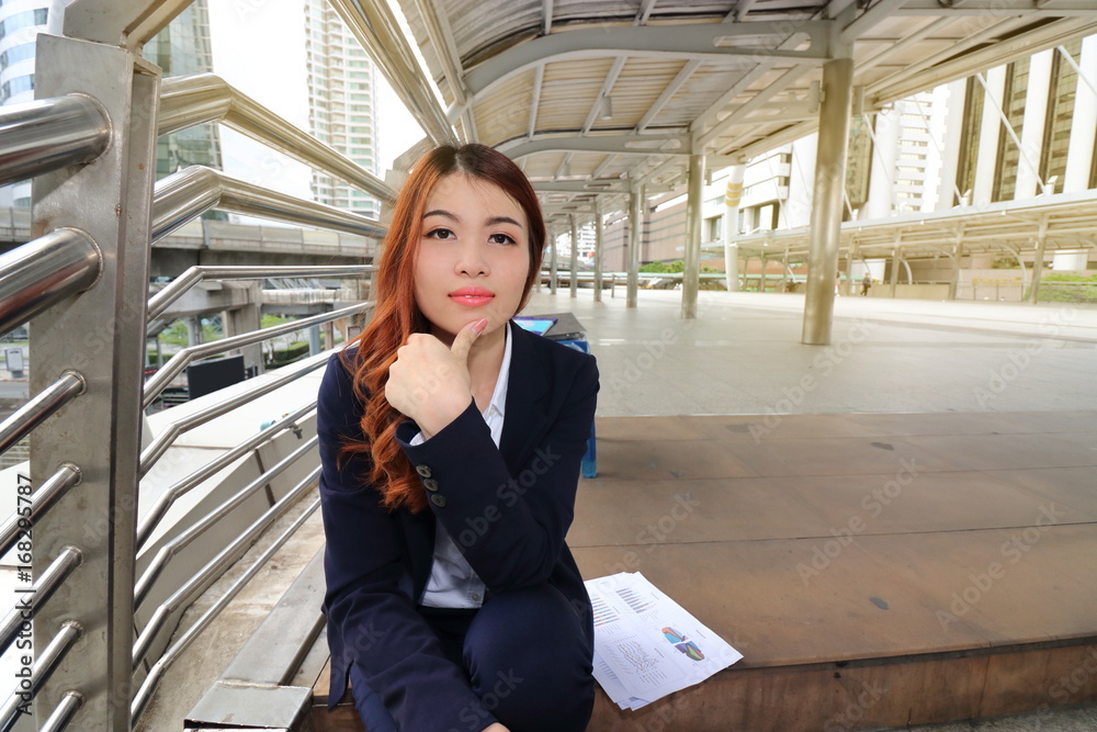 Fototapeta premium Portrait of attractive young Asian businesswoman sitting on stair way and looking on camera in urban building background.