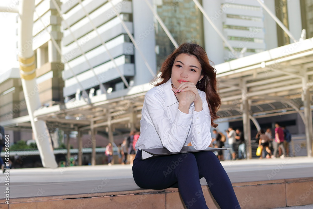 Portrait of leader young Asian business woman thinking and sitting on stairway in urban building background.