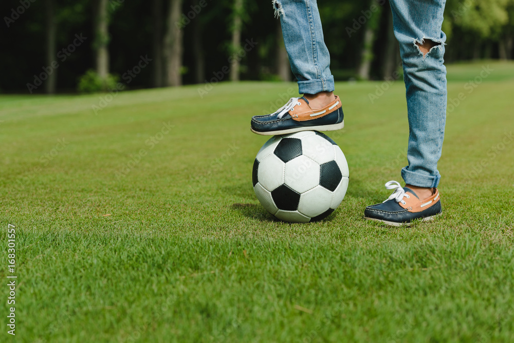 Close-up partial view of kid standing with soccer ball on green meadow
