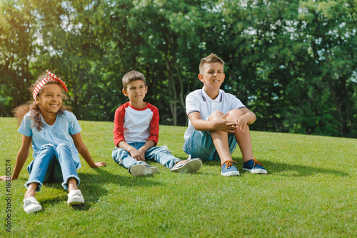 Cute smiling multiethnic kids sitting together on green lawn in park