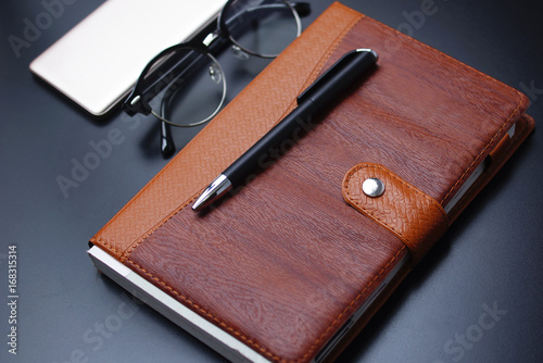 Leather Management Diary , Ball Pen , Black Eye Glasses , And Smartphone Over Black Background