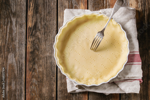 Dough for baking quiche tart in ceramic baking form ready for bake on kitchen towel over old plank wooden background. Flat lay with copy space