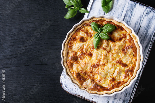 Baked homemade quiche pie in white ceramic form served with fresh greens on white serving board on black wooden burned background. Flat lay with copy space photo