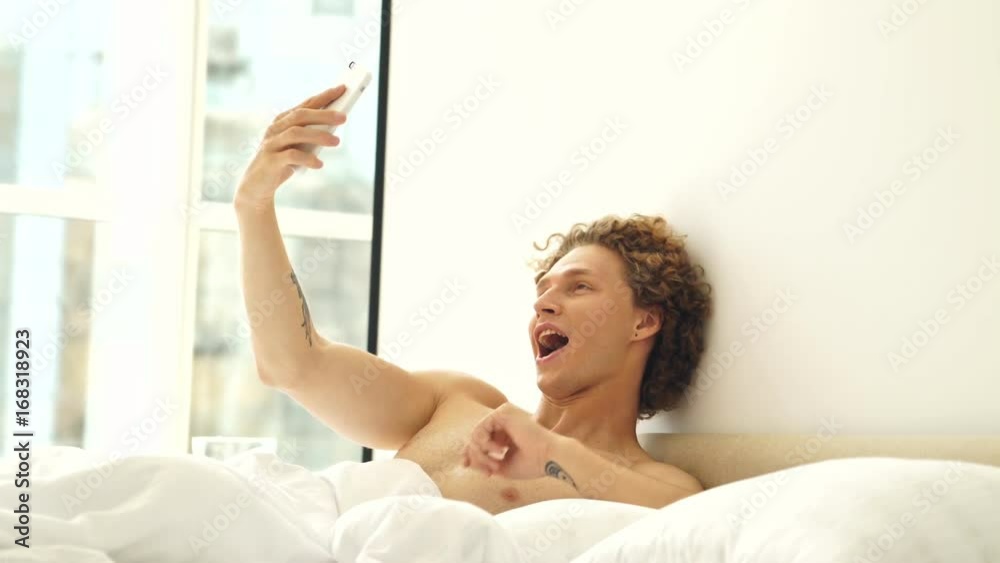 Young handsome man lying in bed at home and making selfie with different emotions on smartphone Young handsome man lying in bed at home and making selfie with different emotions on smartphone
