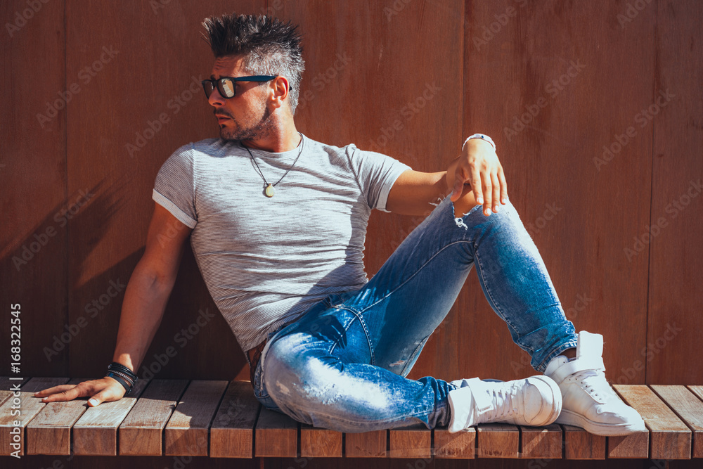 Portrait of handsome young man in casual clothing with sunglasses sitting on wood bench, looking away to a side. Color Toned Image. Concept urban man. Portrait of handsome young man in casual clothing with sunglasses sitting on wood bench, looking away to a side. Color Toned Image. Concept urban man.