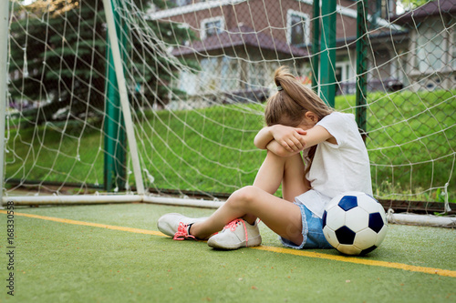 Crying ltittle girl who is playing football, hiding her face while being upset to miss goal in gates. Pretty kid wearing white t-shirt, jean shorts and sport shoes, playing football. Children, sport